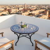 Cobalt blue and white zellige mosaic table on a white Moroccan rooftop terrace with silver teapot blue tea glasses jasmine flowers and medina cityscape view
